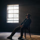 Person sitting on a chair in a dimly lit room with a barred window.